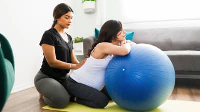Pregnant woman leaning on exercise ball with trainer helping stretch the lower back.